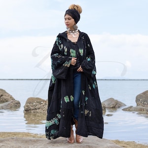 Op de afbeelding: Een lange, zwarte jas met capuchon met een patchwork-ontwerp en groene en blauwe accenten. De jas wordt gedragen over een blauwe spijkerbroek en bruine laarzen. De achtergrond toont een watermassa en een bewolkte lucht.