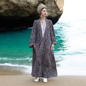 May include: A woman wearing a long, black and white leopard print robe with a light brown headscarf. She is standing on a sandy beach with turquoise water in the background.