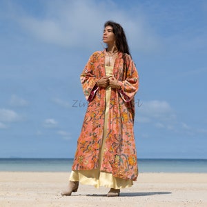May include: A long, pink floral print kimono with a yellow undergarment. The kimono has a wide, flowing silhouette and features a floral pattern in shades of pink, orange, and green. The model is standing on a sandy beach with a blue sky and ocean in the background.