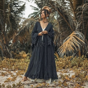 May include: A long, dark gray dress with a flowing silhouette. The dress features a deep V-neck and long sleeves with ruffled detailing. The model is wearing a white slip underneath. The background includes palm trees and beach vegetation.