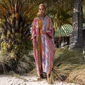 May include: A woman wearing a long, pink and blue patterned kimono with a yellow dress underneath. She is standing in front of a palm tree and has a head scarf on.