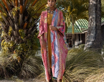 May include: A woman wearing a long, pink and blue patterned kimono with a yellow dress underneath. She is standing in front of a palm tree and has a head scarf on.