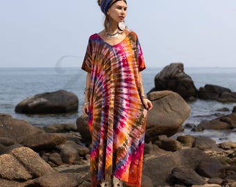 May include: A colorful tie-dye maxi dress with a V-neck. The dress features a vibrant mix of orange, pink, purple, and brown hues. The model is standing on rocks near the ocean, wearing white boots and a blue headscarf.