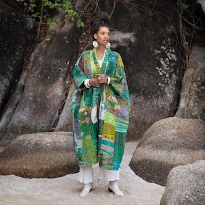 May include: A long, patchwork kimono in shades of green, yellow, and purple. The kimono features a variety of fabric patterns and textures. The person is wearing white pants and boots. The background includes large rocks and foliage.