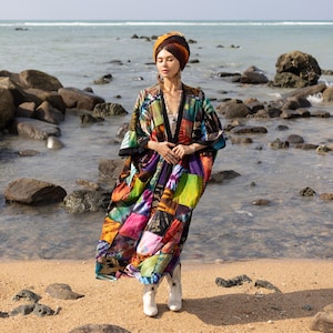 May include: A woman wearing a colorful patchwork kimono-style robe with a head scarf and white boots, standing on a beach with rocks and water in the background.