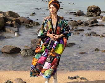 May include: A woman wearing a colorful patchwork kimono-style robe with a head scarf and white boots, standing on a beach with rocks and water in the background.