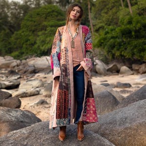 May include: A long, patchwork coat in various colors, including pink, blue, and brown, worn over a peach-colored top and blue jeans. The coat has a flowing, open design and is paired with brown boots. A necklace is visible.
