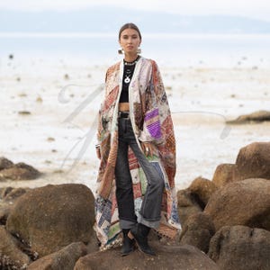 May include: A long patchwork kimono in shades of cream, red, purple, and blue. The kimono is worn over a black top and dark wash jeans. The model is standing on a rock near the ocean.