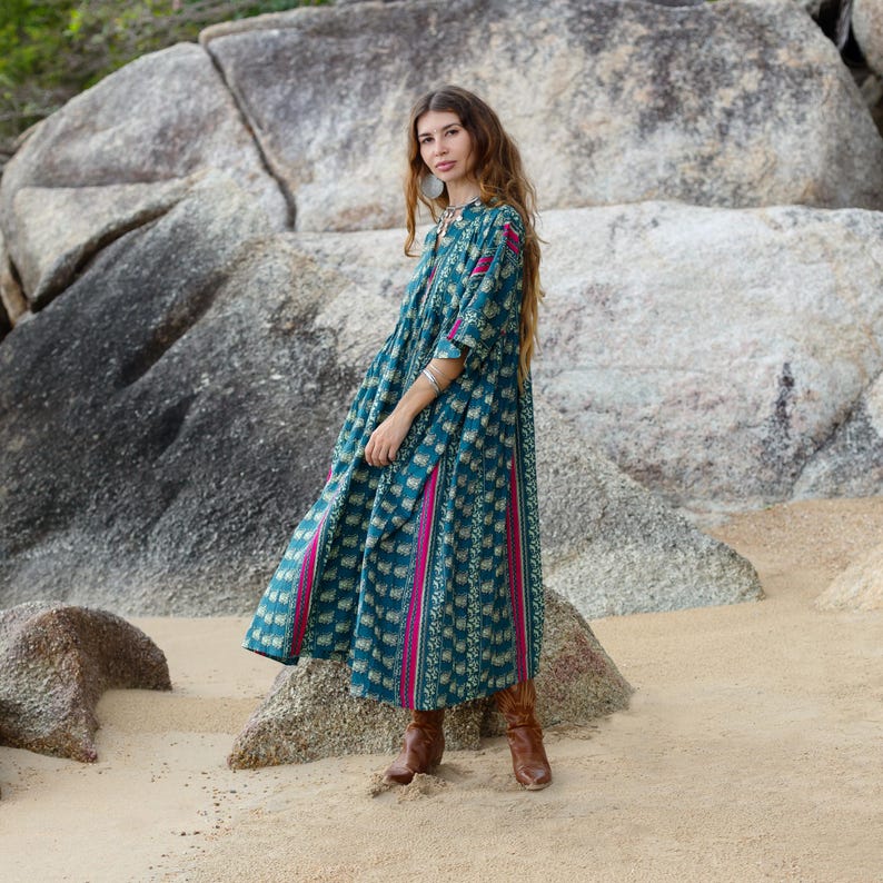 4. Side profile of model wearing the Cora Midi Dress in Peacock Rose on a coastal rocky beach. The lightweight cotton boho dress features a relaxed babydoll silhouette, wide sleeves, deep pockets, and flowing teal patterned fabric accented with bold magenta panels. The airy oversized drape creates graceful movement perfect for festival styling, resort wear, and artisan everyday dressing.
