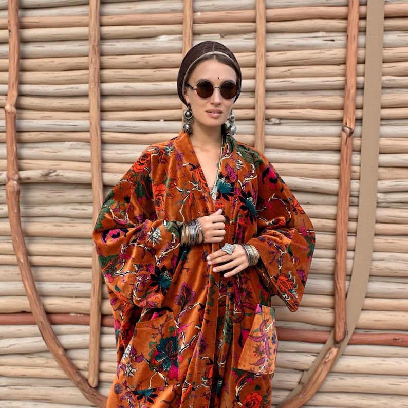 Close-up portrait of the Amber Flora Harper Jade Velvet Kimono Robe, photographed against a rustic woven wooden wall. The handcrafted velvet duster features jewel-toned floral motifs across a warm sienna ground, wide flowing sleeves, and visible patch pockets. Styled with silver bangles, statement earrings, and a hair wrap, this bohemian robe radiates artisanal elegance. Perfect as a plus size fall festival coat, holiday party duster, or timeless boho gift.