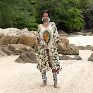 May include: A woman wearing a long, white, tie-dye dress with green and orange accents. She is standing on a sandy beach with large rocks in the background. She is wearing a silver necklace and large silver earrings.