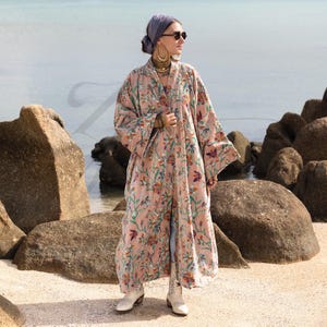 May include: A long, open-front robe in a soft pink hue, adorned with a floral pattern in shades of green, orange, and purple. The robe features wide sleeves and a flowing silhouette. The model is wearing white boots and a gray head covering.
