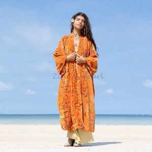 May include: A long orange floral print kimono with a light brown belt. The kimono is worn by a person standing on a sandy beach with a blue sky and ocean in the background.