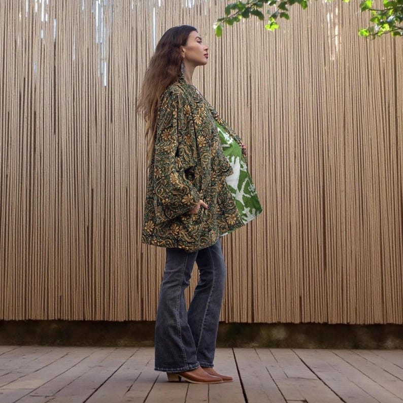 Profile view showing the draped silhouette of the emerald floral velvet kimono jacket with contrasting green block-printed lining, styled with flared jeans and silver jewelry.