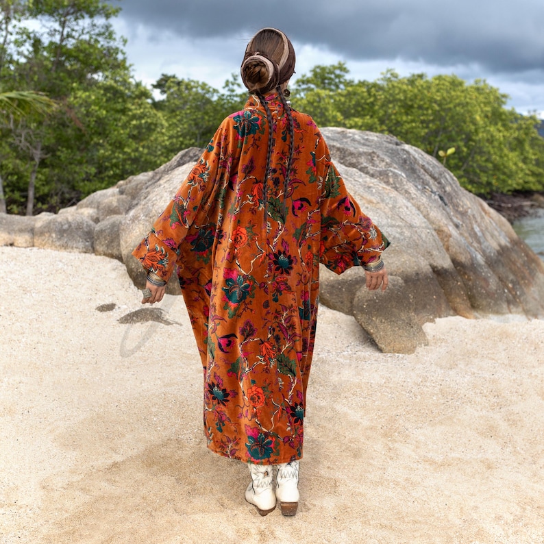 Back view of the Amber Flora Harper Jade Velvet Kimono Robe by ZinniaPearl, showing the full oversized silhouette flowing against rugged rock formations and greenery. The handcrafted velvet coat features an autumn-inspired sienna ground with intricate floral motifs in magenta, teal, and emerald. With wide sleeves, a floor-length cut, and deep pockets, this plus size bohemian duster radiates dramatic presence, ideal for fall ceremonies, holiday gatherings, and slow fashion styling.