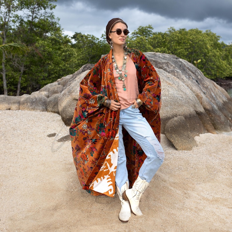 Model wearing the Amber Flora Harper Jade Velvet Kimono Robe by ZinniaPearl, standing confidently against large stone rocks and lush green trees under a moody sky. The plus size boho duster is crafted from cotton velvet in warm sienna tones with jewel-toned florals in teal, magenta, and emerald. Featuring wide sleeves, deep pockets, and an oversized fit, this handcrafted robe flows beautifully, styled with distressed jeans, silver jewelry, and white boots.