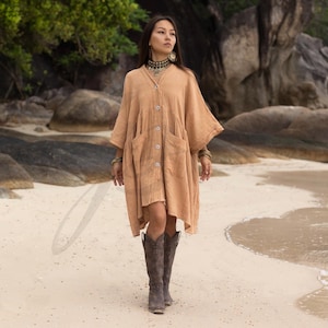 May include: A woman wearing a long, light brown, button-down shirt dress with pockets and brown cowboy boots. She is standing on a sandy beach.