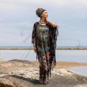 May include: A tie-dye caftan dress with fringe trim. The dress features a circular design in the center with shades of green, purple, and blue. The dress is knee-length and has long fringe along the sleeves and hemline. The model is wearing brown boots.