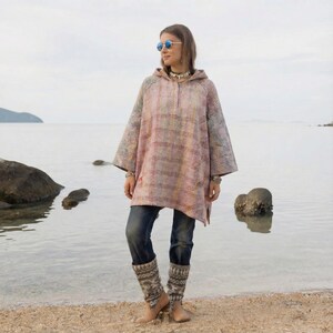 Pode incluir: Um poncho com capuz em patchwork com tons de rosa, vermelho e azul. O poncho folgado é usado com jeans azuis e polainas estampadas. O fundo mostra uma praia e o mar.