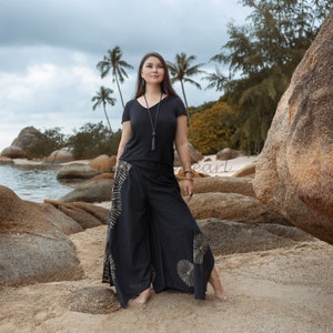 Pode incluir: Uma mulher vestindo uma camiseta preta e calças largas pretas com um padrão tie-dye está em uma praia de areia com grandes rochas e palmeiras ao fundo.