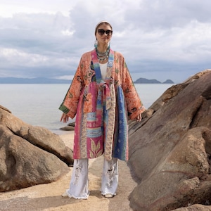 Puede incluir: Una colorida bata estilo kimono patchwork con cinturón anudado, con tonos naranja, rosa, azul y verde. La bata se usa con vaqueros desgastados de color claro y accesorios. El fondo muestra una escena de playa con rocas y el océano.