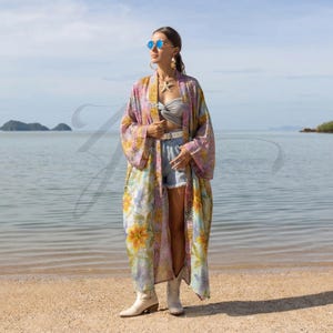 Puede incluir: Un kimono largo y fluido en tonos rosas, amarillos y azules, con un estampado floral. El kimono se lleva sobre un top gris, unos shorts vaqueros y botas blancas. La modelo lleva gafas de sol y un collar de estrella de mar.