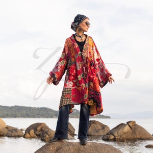May include: A patchwork kimono-style jacket in shades of red, orange, and black. The jacket features a variety of floral and geometric patterns. The person is wearing dark blue jeans, a black top, and a patterned headscarf. The background shows a body of water and rocks.