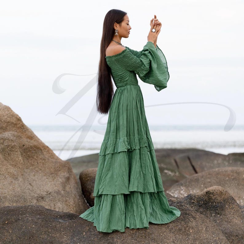 This off-shoulder green cotton maxi dress is perfect for bohemian weddings, outdoor elopements, or forest gatherings. Lucia by ZinniaPearl features a flared long sleeve, stretchy smocked bodice, and tiered flowy skirt made of raw natural cotton. Elderfern Green evokes woodland serenity and botanical grace, making this handmade gown a top choice for free-spirited brides, maternity shoots, and slow fashion lovers.