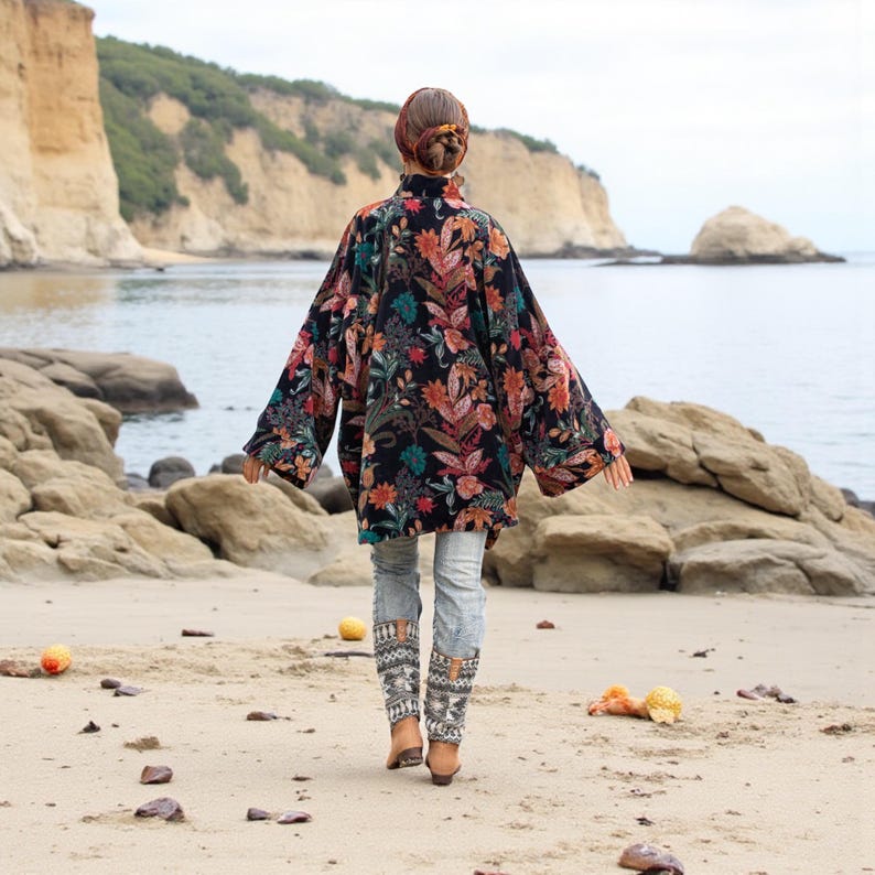 Back view of the Mei Velvet Kimono Duster in Nocturne Flora, highlighting the dramatic drape and oversized sleeves. The deep black velvet base blooms with botanical patterns in warm and cool jewel tones. The model walks barefoot among rocks by the sea, surrounded by soft coastal light and sand, embodying the effortless grace, flow, and confidence of ZinniaPearl’s bohemian aesthetic.