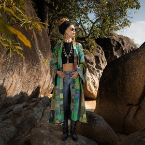 May include: A woman wearing a green and blue patchwork kimono with a black top and jeans. She is wearing a silver necklace and sunglasses. The woman is standing in front of large rocks.