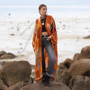 May include: An orange patchwork kimono with long sleeves. The kimono features a variety of patterns and colors, including red, yellow, and brown. The model is wearing a black top, gray jeans, and black boots. The background is a beach scene.