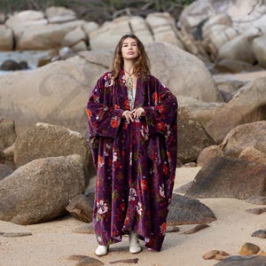 Pode incluir: Um casaco duster comprido e largo de veludo roxo com um padrão floral vibrante de flores vermelhas, brancas e verdes. A peça tem mangas largas a todo o comprimento e um design frontal aberto, usado sobre um top claro e botas brancas.