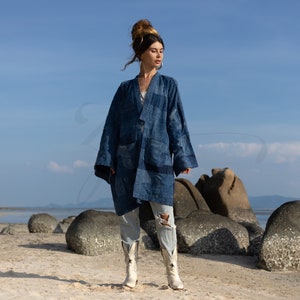 Op de afbeelding: Een vrouw draagt een blauw denim jack met een patchwork ontwerp, gescheurde jeans en witte cowboylaarzen. Ze staat op een strand met grote rotsen op de achtergrond.