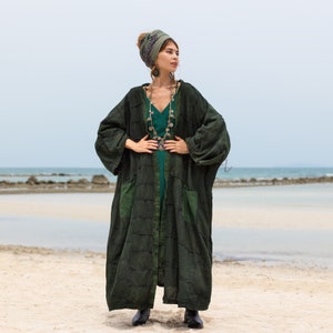 Könnte beinhalten: Dunkelgrüner Patchwork-Robe mit großen Taschen und langem, fließendem Design. Die Robe wird über einem türkisfarbenen Kleid getragen und mit einem gemusterten Kopftuch und einer silbernen Halskette kombiniert. Im Hintergrund ein Strand und das Meer.