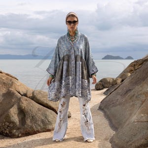 May include: A woman in a patterned blue, grey, and brown tunic with wide sleeves and distressed white jeans. She wears sunglasses, a headband, and turquoise jewellery. The background shows a beach and the sea.