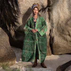 May include: A long, flowing green kimono with a paisley pattern. The kimono is worn over a dark top and blue jeans. The model is wearing brown boots and a blue and yellow head wrap. The background features large rock formations.