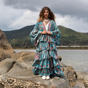 May include: A teal, tiered maxi dress with a floral pattern and bell sleeves. The dress features red and pink accents and is worn with white shoes. The model is standing on a rock near the water.