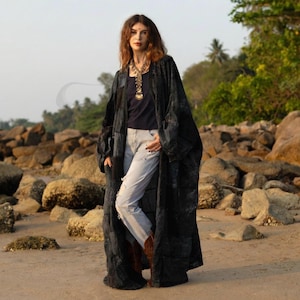 Może przedstawiać: Długa, czarna szata w stylu kimono z patchworkiem. Szata jest noszona na ciemnoniebieskim topie i jasnoniebieskich dżinsach. Osoba nosi złoty naszyjnik i brązowe buty. Tło to plaża ze skałami i drzewami.