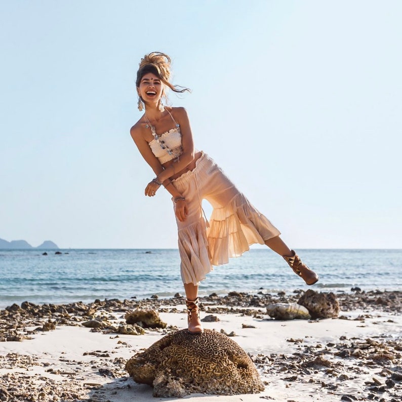 May include: A woman wearing a white crop top and a flowy, tiered skirt stands on a rock formation on a beach. She is smiling and looking at the camera. The beach is sandy with rocks and water in the background.