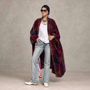 Puede incluir: Un abrigo largo de terciopelo abierto en color burdeos y morado con un estampado floral multicolor, sobre una camiseta blanca sin mangas y vaqueros azules claros, combinado con botines blancos, un pañuelo oscuro y gafas de sol oscuras.