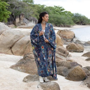 May include: A woman wearing a long, blue, patterned kimono with colourful circles and embroidery. She is standing on a rocky beach with white sand.