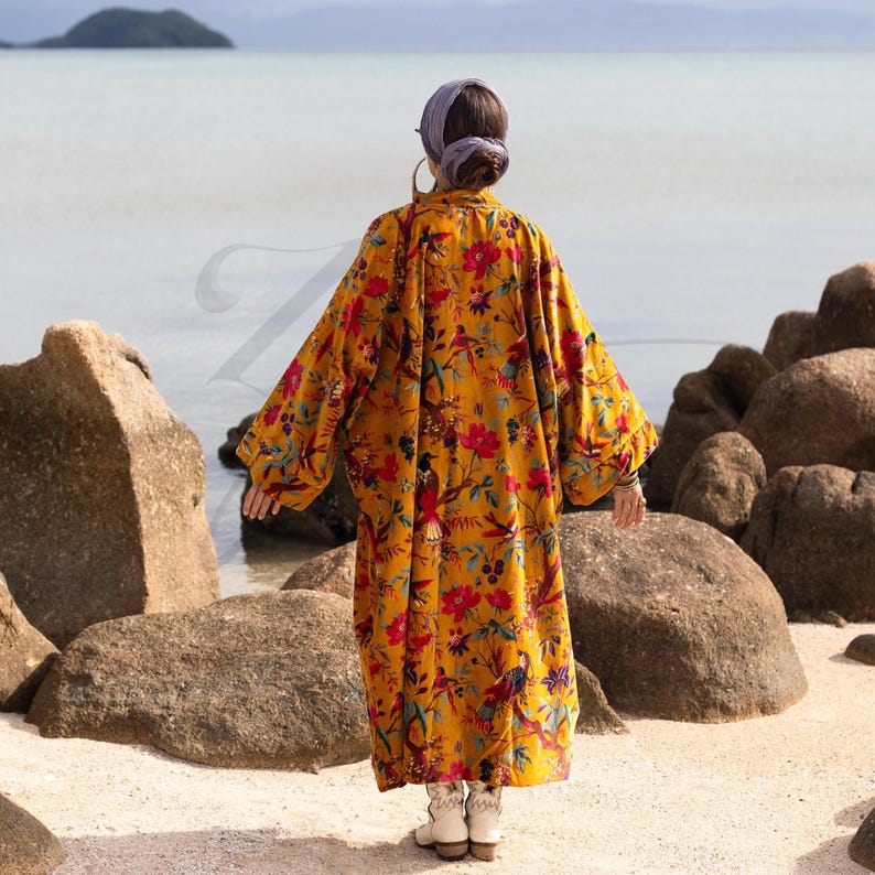 Full-length back view of the Mai velvet kimono in Golden Sunbird print. The oversized duster coat flows to the ankles, displaying the intricate pattern of tropical birds and red blooms on mustard velvet. The wide, drop-shoulder sleeves and generous width emphasize the relaxed, plus-size-friendly fit. The model stands in white western boots on a sandy beach, demonstrating the elegant, sweeping silhouette of this handmade boho robe.