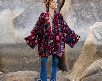 May include: A burgundy velvet kimono with a floral pattern in red, blue, and green. The open-front jacket has wide sleeves and is paired with blue denim jeans with fringed hems. The model is wearing a turban and jewelry.
