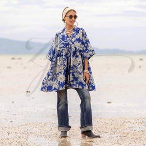 May include: A blue tunic with a white floral design, worn with blue jeans. The tunic features a V-neck and short sleeves. The model is wearing sunglasses, a headband, and jewellery. The background is a beach with a cloudy sky.
