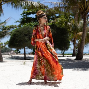 Puede incluir: Una mujer con un vestido largo y fluido de color naranja y verde con estampado tie-dye y un escote en V profundo. Está de pie en una playa de arena blanca con palmeras en el fondo.