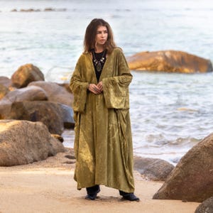 May include: An olive green velvet robe with wide sleeves. The robe is worn over a black top and trousers. The person is standing on a sandy beach with rocks and the sea in the background.