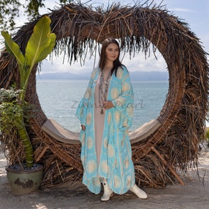 May include: A woman wearing a light blue and white patterned kimono with a long, flowing skirt. She is standing in front of a wicker heart-shaped archway with a view of the ocean in the background.