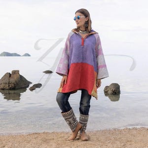 May include: A colorful patchwork tunic with a hood, featuring shades of purple, red, and pink. The tunic has long sleeves and a textured fabric. The person is wearing blue jeans and patterned leg warmers, standing on a sandy beach near the water.