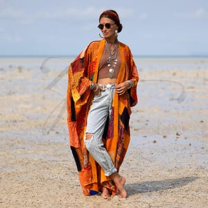 May include: An orange patchwork kimono with long sleeves. The kimono is worn over a brown top and light blue jeans. The outfit is accessorized with silver necklaces, bracelets, and a headband. The background is a beach with a blue sky.