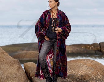 May include: A long, patterned velvet robe in shades of burgundy, blue, and gold. The robe is open, revealing a black top and gray jeans. The model is standing on rocks near the ocean, wearing black boots and layered necklaces. The robe has a bohemian style.